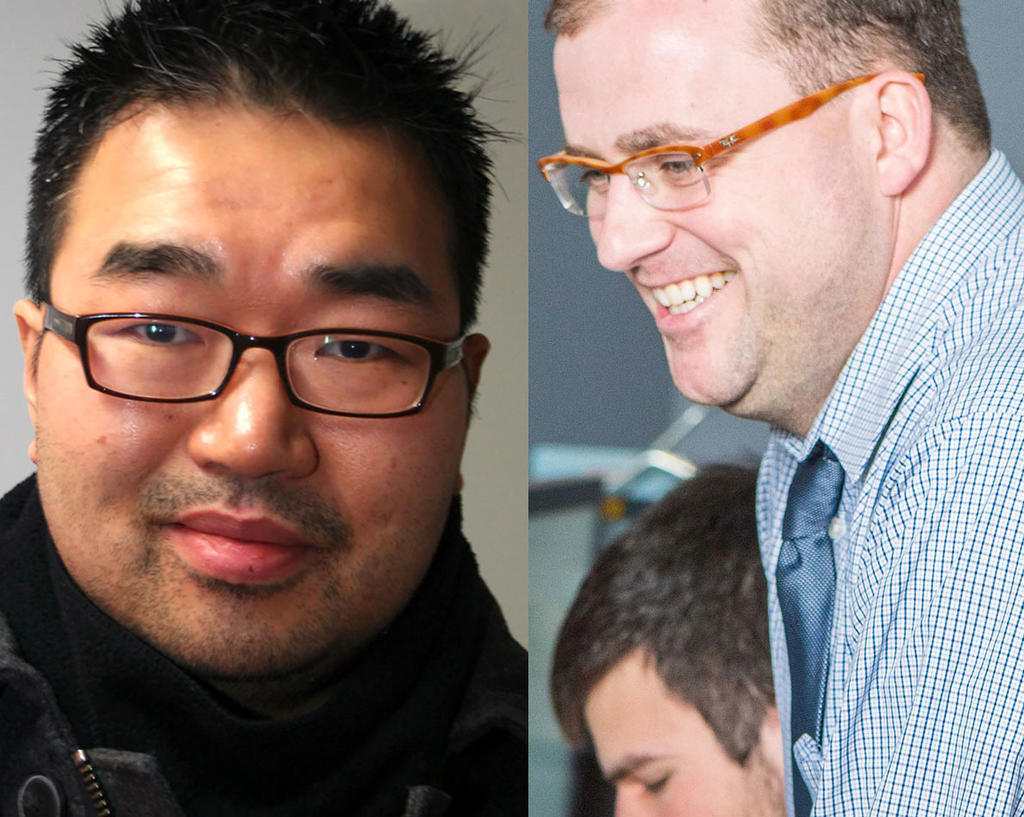 Asian man dressed in black with black glasses and spiked black hair wearing leather jacket and a white man wearing a blue shirt learning over a student at a desk